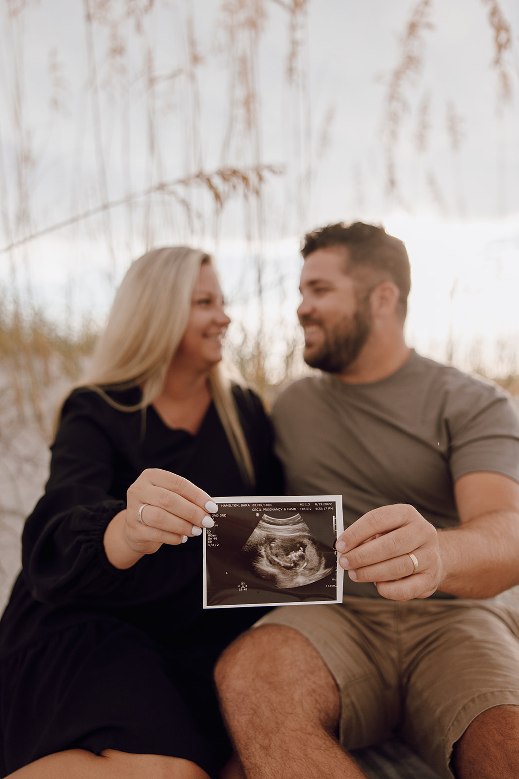 the couple sitting in the sand and holding the baby ultrasound photo