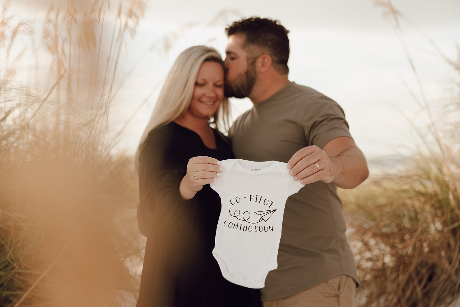 couple standing in the dune grass with a baby onesie for a pregnancy annoucement