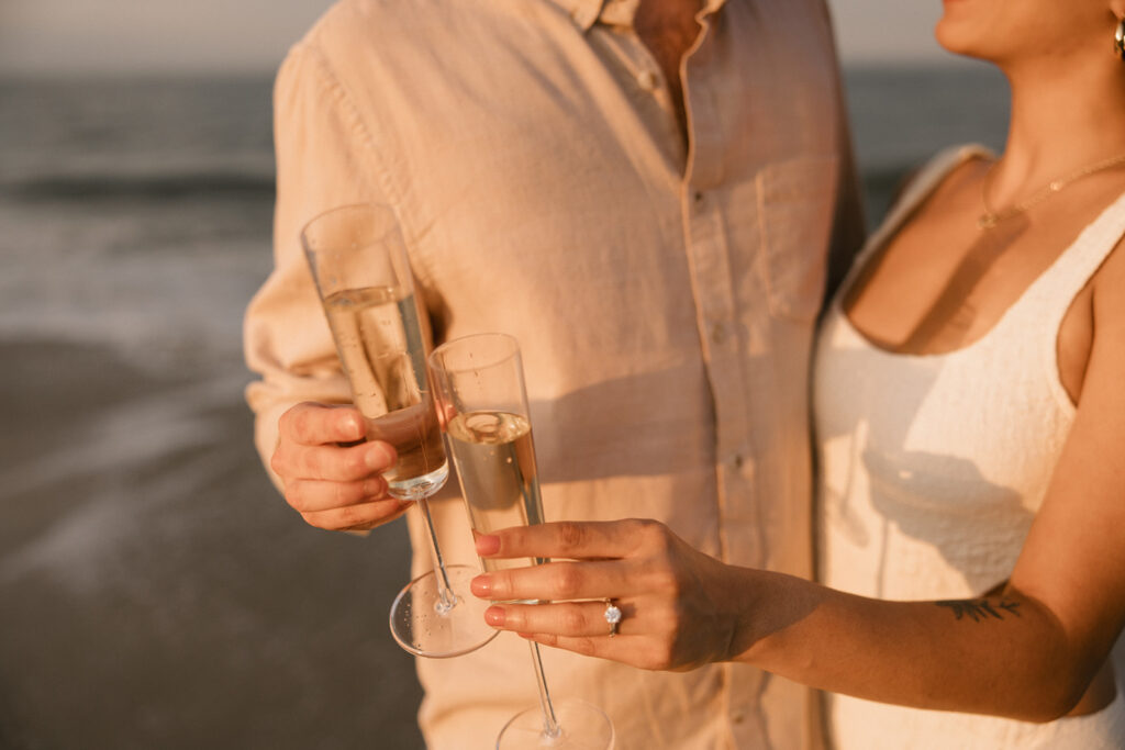 the couple holding their glasses on the beach during their beach engagement photos on the east coast