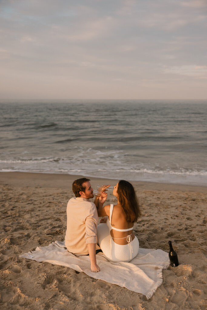 the engaged couple enjoying a drink on a beach blanket for their beach engagement photos