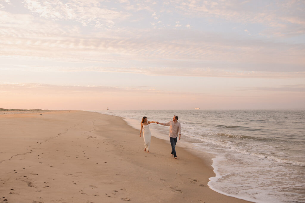 the engaged couple in the distance holding hands as they walk along the sand for their beach engagement photos