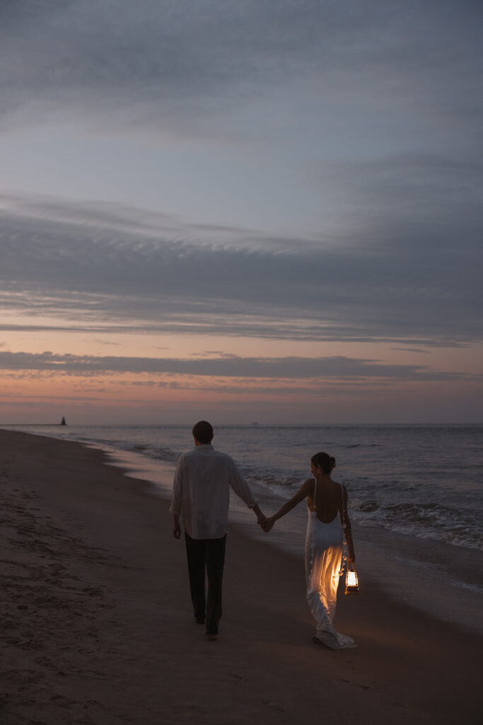 the couple holding hands as they walk down the beach with a lantern for their beach engagement photos