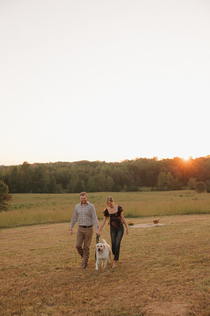 the engaged couple walking with their dog during sunset