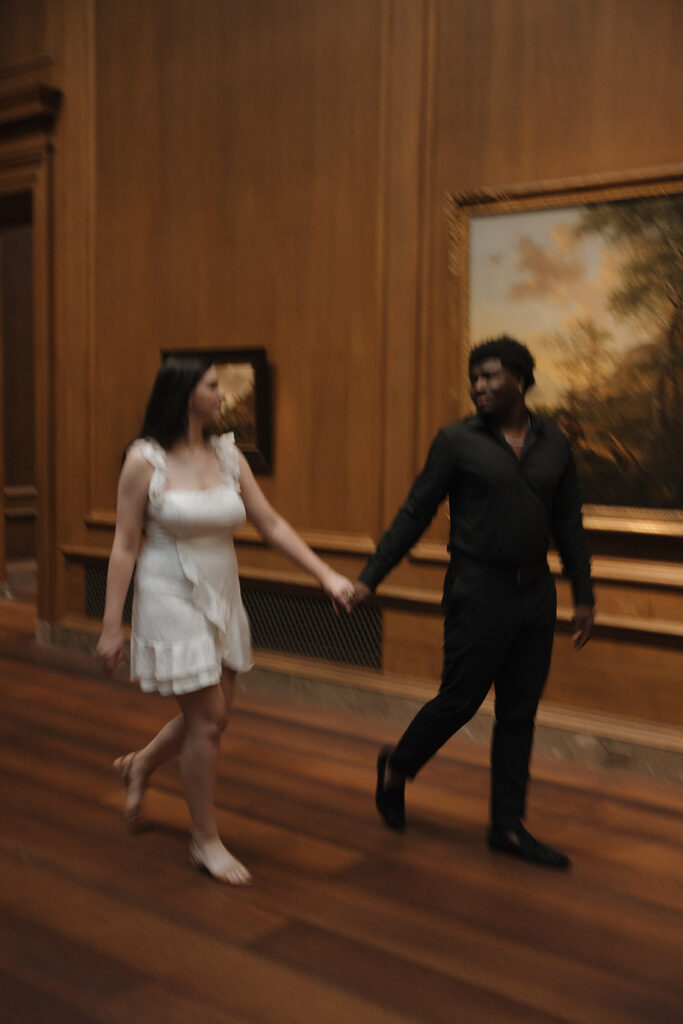 the engaged couple holding hands as they walk through the art gallery 